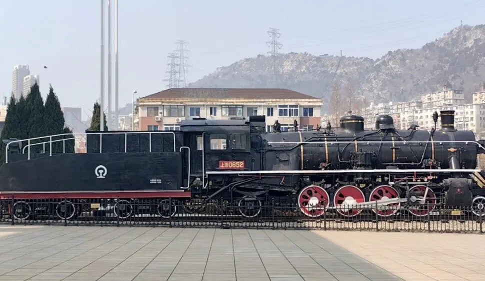 Dalian Modern Museum,Dalian,Liaoning,Chinatravel Historic steam locomotive on display in a public square; black body with red wheels and white piping, mountainous backdrop.