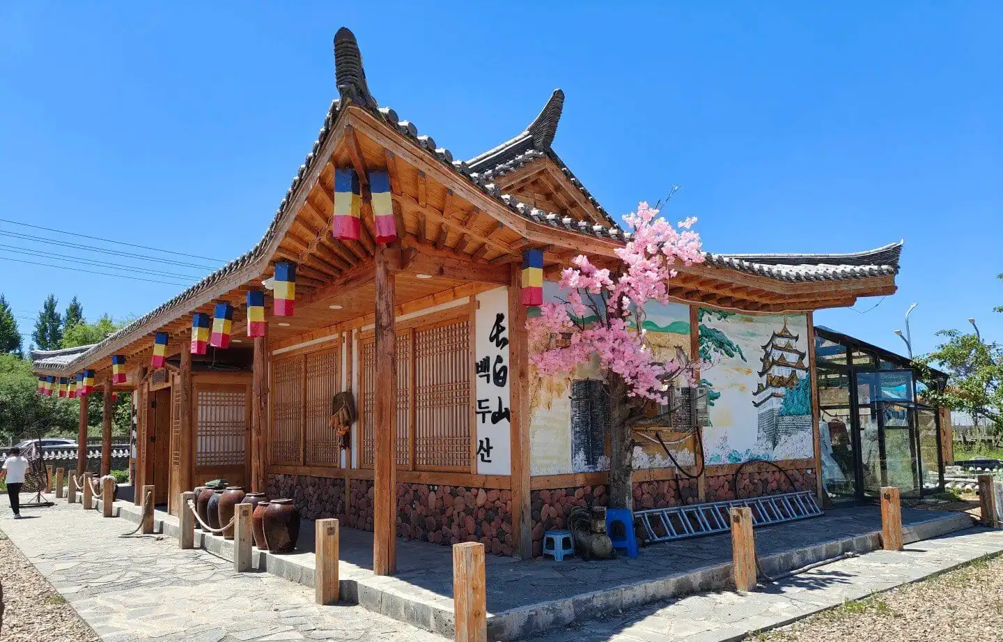 Changbai Korean Folk Village,Baishan,Jilin,Chinatravel Traditional wooden East Asian building with curved tiled roof, colorful banners, and a pink cherry blossom tree in bloom beside a stone path and mural wall.