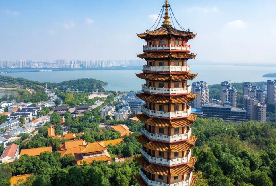 Sheng'an Temple,Yueyang,Hunan,Chinaexplorertour Tall multi-tier Chinese pagoda with orange-tiled roofs sits atop a hill, overlooking a river, trees, and a cityscape of modern apartments and traditional buildings in the distance.