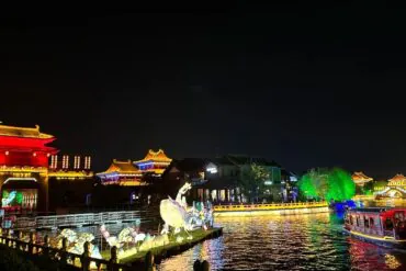 Millennium City Park,Kaifeng,Henan,Chinatravel Night view of a lit riverside with traditional East Asian pagodas, red and gold buildings, and colorful lamp-lit structures reflecting on the water; a boat glides along the river.