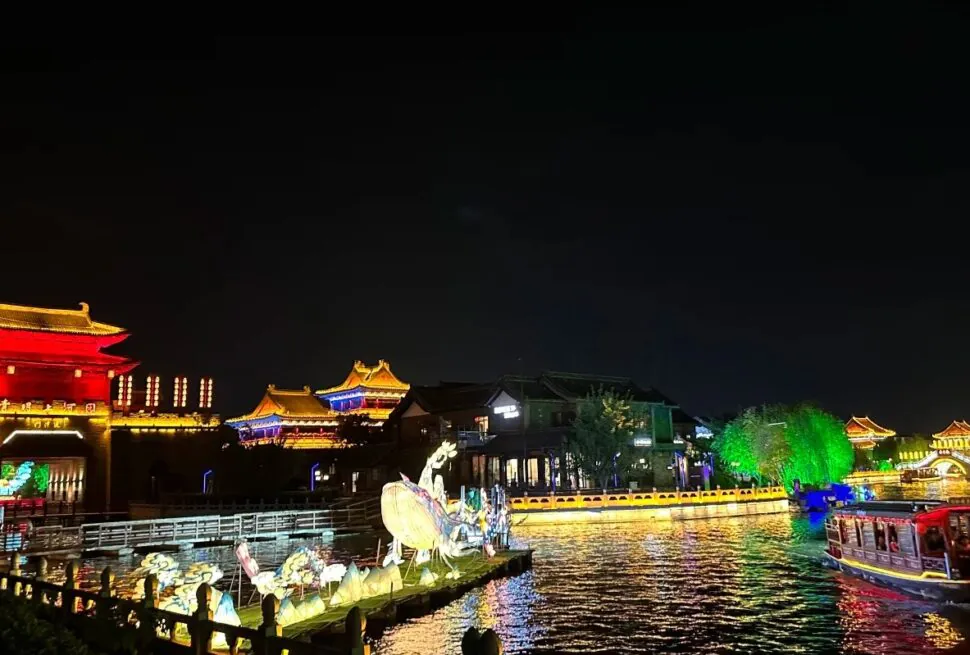 Millennium City Park,Kaifeng,Henan,Chinatravel Night view of a lit riverside with traditional East Asian pagodas, red and gold buildings, and colorful lamp-lit structures reflecting on the water; a boat glides along the river.