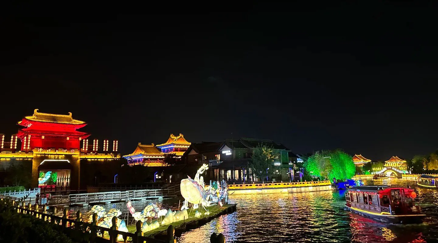 Millennium City Park,Kaifeng,Henan,Chinatravel Night view of a lit riverside with traditional East Asian pagodas, red and gold buildings, and colorful lamp-lit structures reflecting on the water; a boat glides along the river.