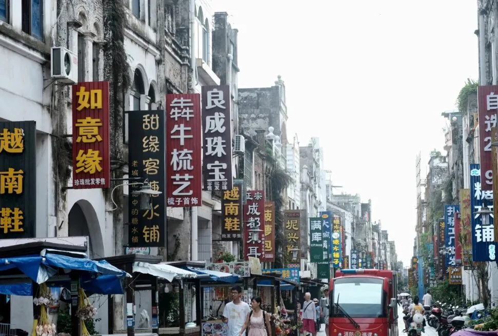 Beihai Old Street,Beiahi,Guangxi,Chinaexplorertour Busy street in China lined with tall vertical signs in Chinese characters and market stalls on both sides, with pedestrians and a red bus in the distance.