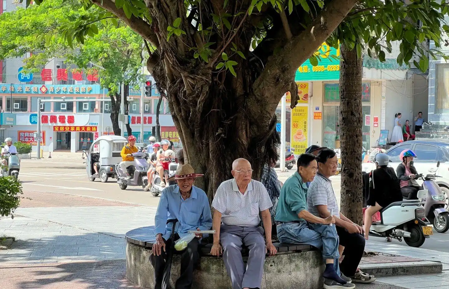 Jiaji Old Town,Qionghai,Hainan,Chinaexplorertour Elderly men sitting on a circular bench under a large tree in a busy urban street scene with scooters and storefronts in the background.