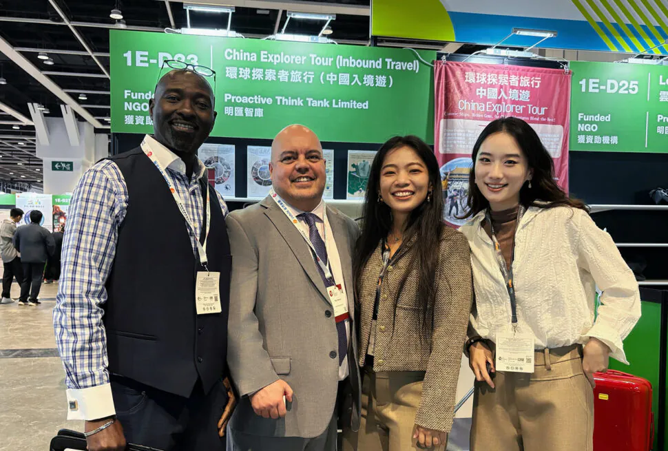 Four professionals pose for a group photo at a trade show booth with green signage overhead, smiling and wearing lanyards.