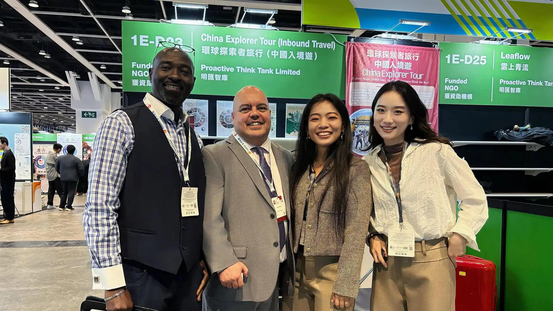 Four professionals pose for a group photo at a trade show booth with green signage overhead, smiling and wearing lanyards.
