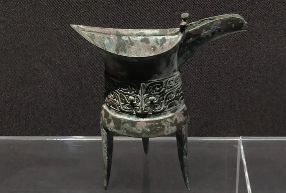 Zhengzhou Museum,Zhengzhou,Henan,Chinaexplorertour Ancient bronze ritual vessel with a long curved spout, standing on three legs, featuring ornate scrolling decorations around the middle.