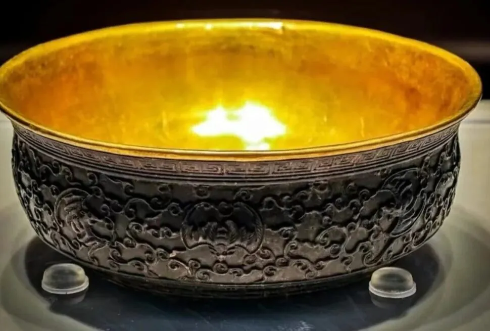 Hainan Museum,Haikou,Hainan,Chinaexplorertour Ornate dark bronze bowl with a gold interior on display, resting on small clear stands.