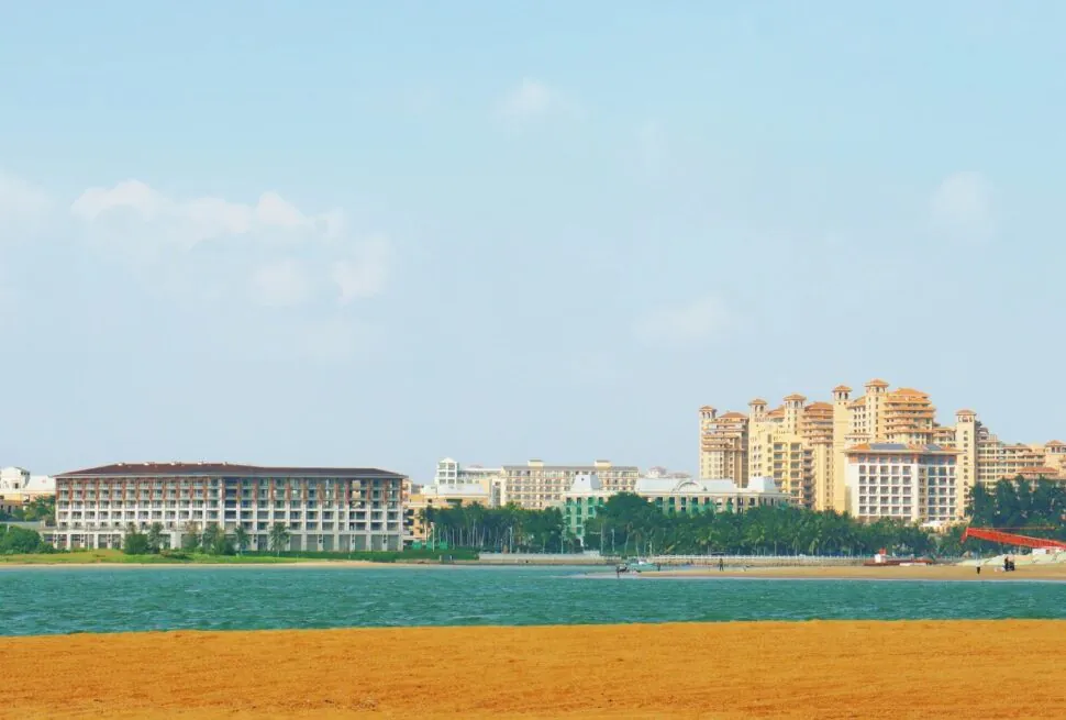 Bo’ao Jade Belt Beach,Qionghai,Hainan,Chinaexplorertour Beach scene with orange sand in the foreground, blue water, and cluster of resort hotels along the shore under a clear sky.