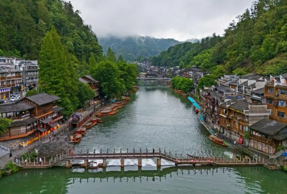 Qinglong Ancient Bridge & Miao Architecture,Fenghuang,Hunan,Chinaexplorertour River town along a calm river with boats docking at the banks and a stone bridge in the foreground. Lush green hills surround traditional multi-story buildings on both sides.