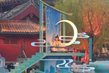 Kaifeng Mansion,Kaifeng,Henan,Chinaexplorertour Performer in traditional clothing sits on a turquoise stage with decorative neon shapes and a large illuminated crescent moon sculpture in a temple courtyard setting.