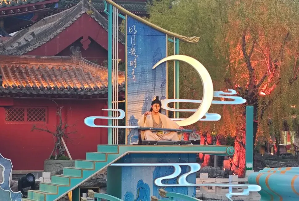 Kaifeng Mansion,Kaifeng,Henan,Chinaexplorertour Performer in traditional clothing sits on a turquoise stage with decorative neon shapes and a large illuminated crescent moon sculpture in a temple courtyard setting.