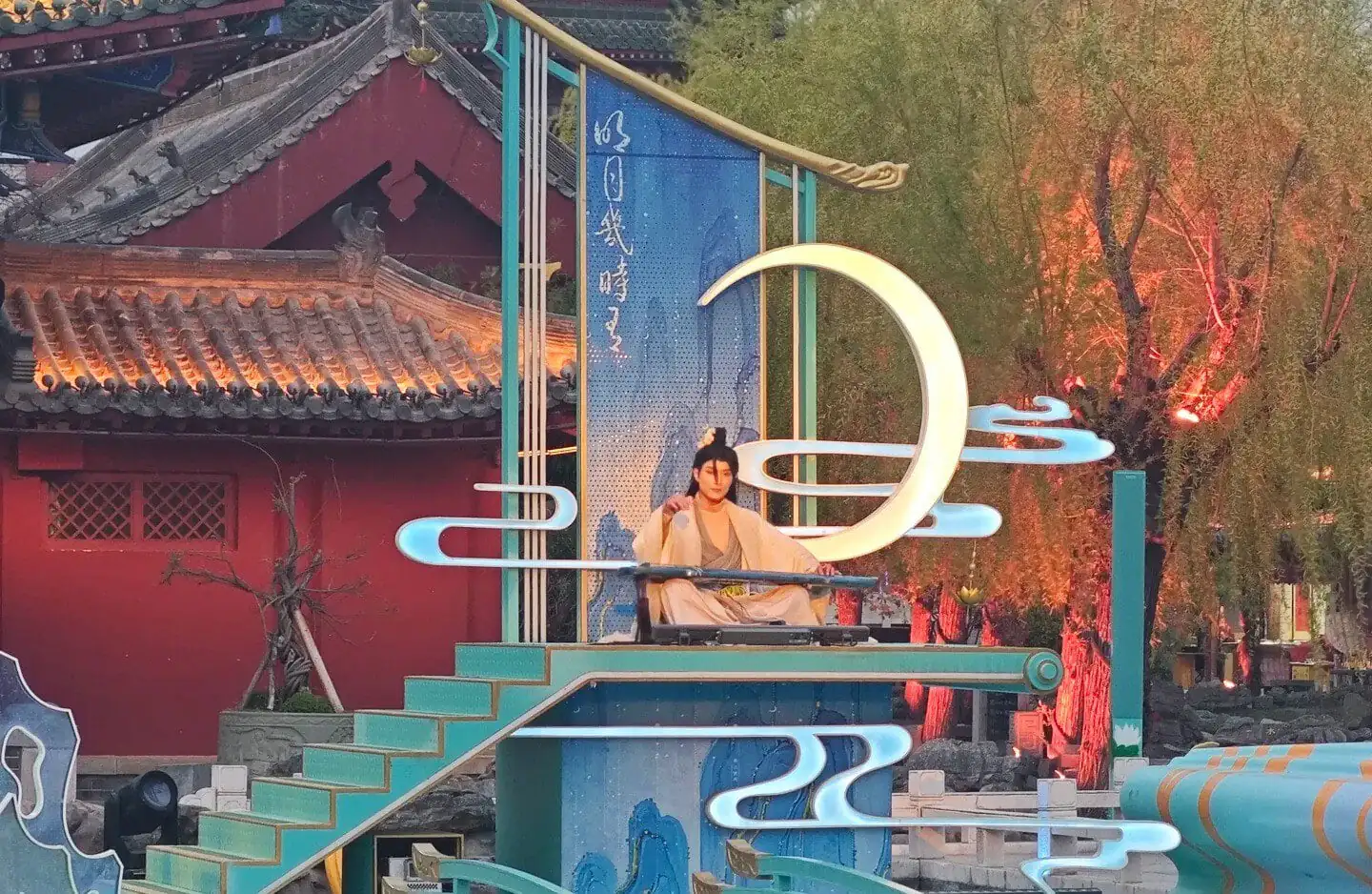 Kaifeng Mansion,Kaifeng,Henan,Chinaexplorertour Performer in traditional clothing sits on a turquoise stage with decorative neon shapes and a large illuminated crescent moon sculpture in a temple courtyard setting.