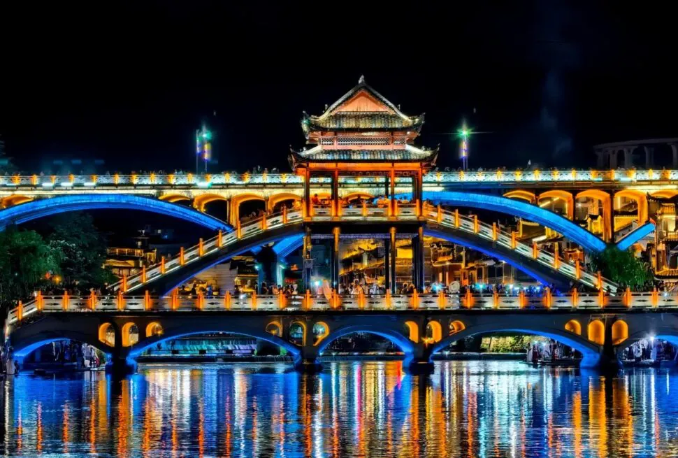 Fenghuang Ancient Town,Fenghuang,Hunan,Chinaexplorertour Night photograph of a pagoda-style bridge lit in blue and orange, spanning a calm river with colorful reflections and a central pavilion.