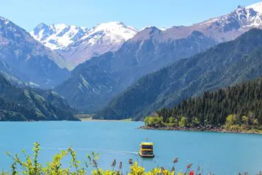 Heavenly Lake of Tianshan,Urumqi,Xinjiang,ChinaexplorertourTurquoise lake with a yellow-and-blue boat and forested mountains with snow-capped peaks in the background