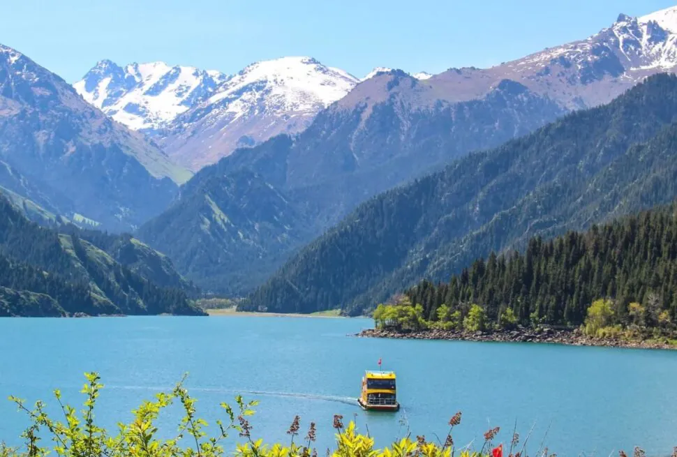Heavenly Lake of Tianshan,Urumqi,Xinjiang,ChinaexplorertourTurquoise lake with a yellow-and-blue boat and forested mountains with snow-capped peaks in the background