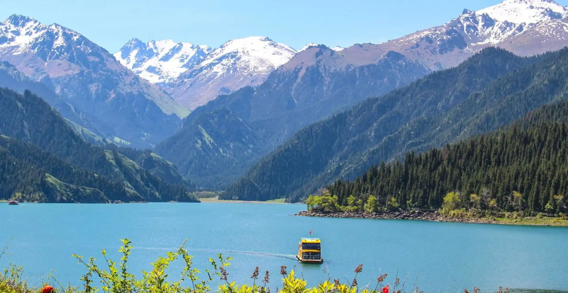 Heavenly Lake of Tianshan,Urumqi,Xinjiang,ChinaexplorertourTurquoise lake with a yellow-and-blue boat and forested mountains with snow-capped peaks in the background