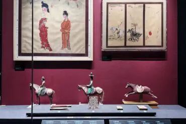 Xinjiang Regional Museum,Chinaexplorertour Museum display: two framed Asian paintings on a burgundy wall and three horse figurines on a blue platform below.