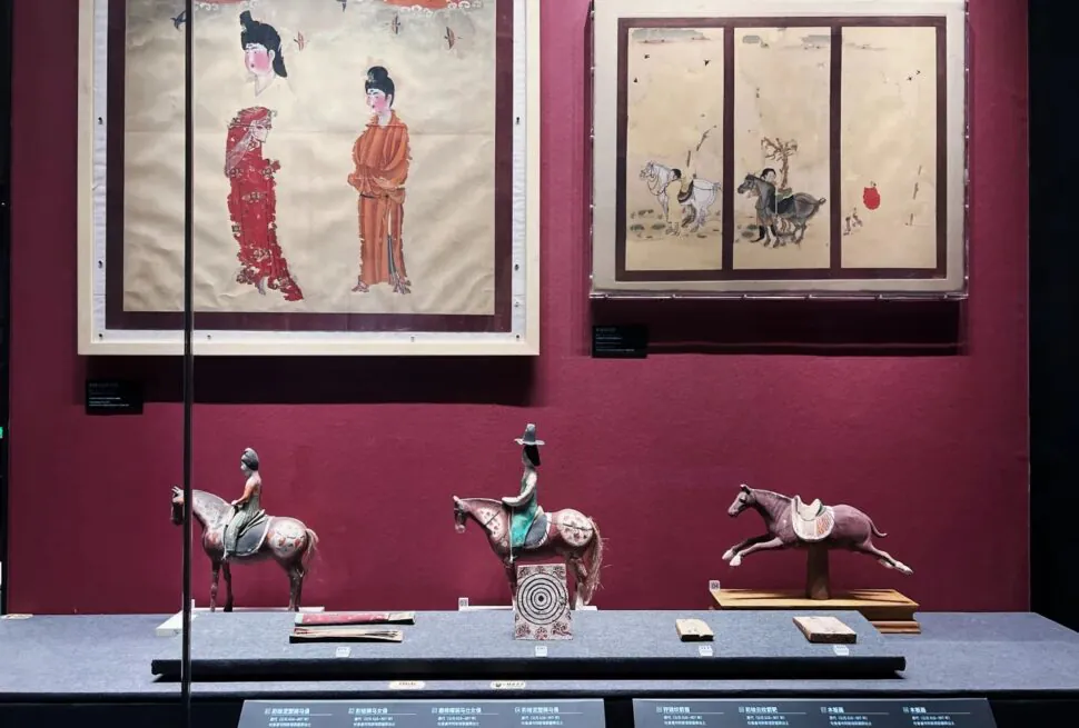Xinjiang Regional Museum,Chinaexplorertour Museum display: two framed Asian paintings on a burgundy wall and three horse figurines on a blue platform below.