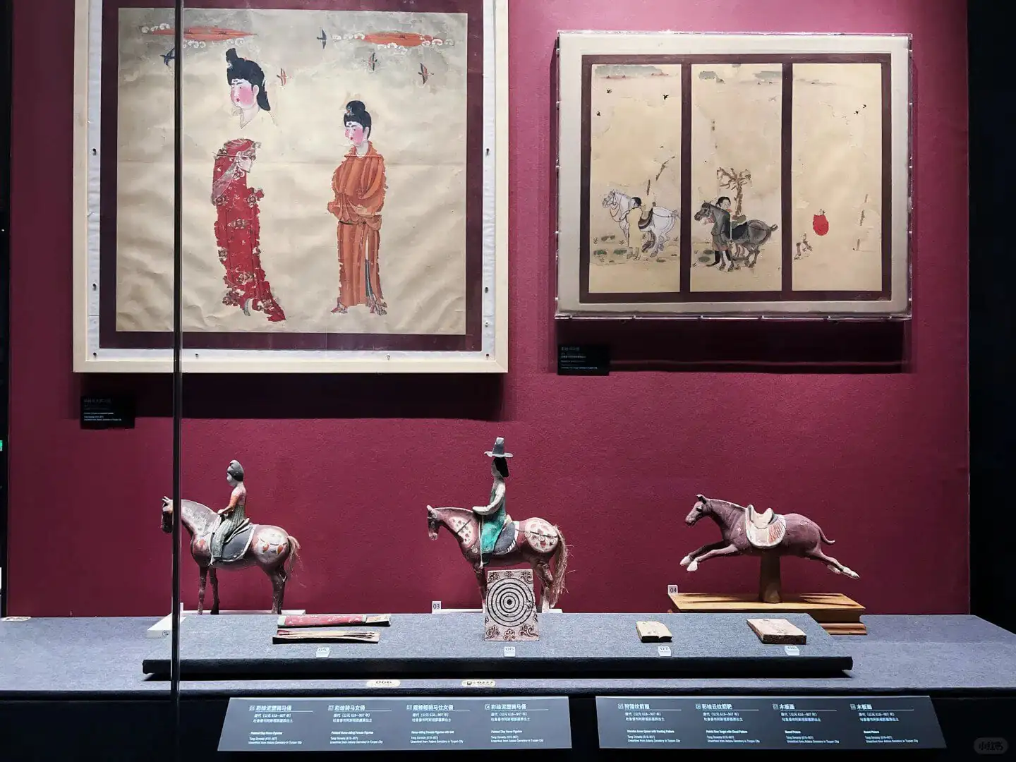 Xinjiang Regional Museum,Chinaexplorertour Museum display: two framed Asian paintings on a burgundy wall and three horse figurines on a blue platform below.
