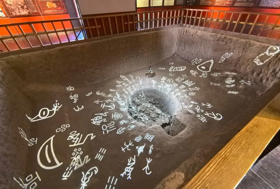 Yin Ruins (Yinxu, UNESCO),Anyang,Chinaexplorertour Indoor museum exhibit: a sunken rectangular pit with a central rock-filled hole, surrounded by white glyphs on the floor and a railing along the edge.