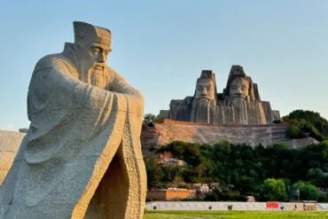 Yellow River Scenic Area,Zhengzhou,Henan,Chinaexplorertour Yellow River Scenic Area,Zhengzhou,Henan,ChinaexplorertourA large seated stone statue in a park with a multi-face rock sculpture carved into a hill in the background, surrounded by trees and landscaping.