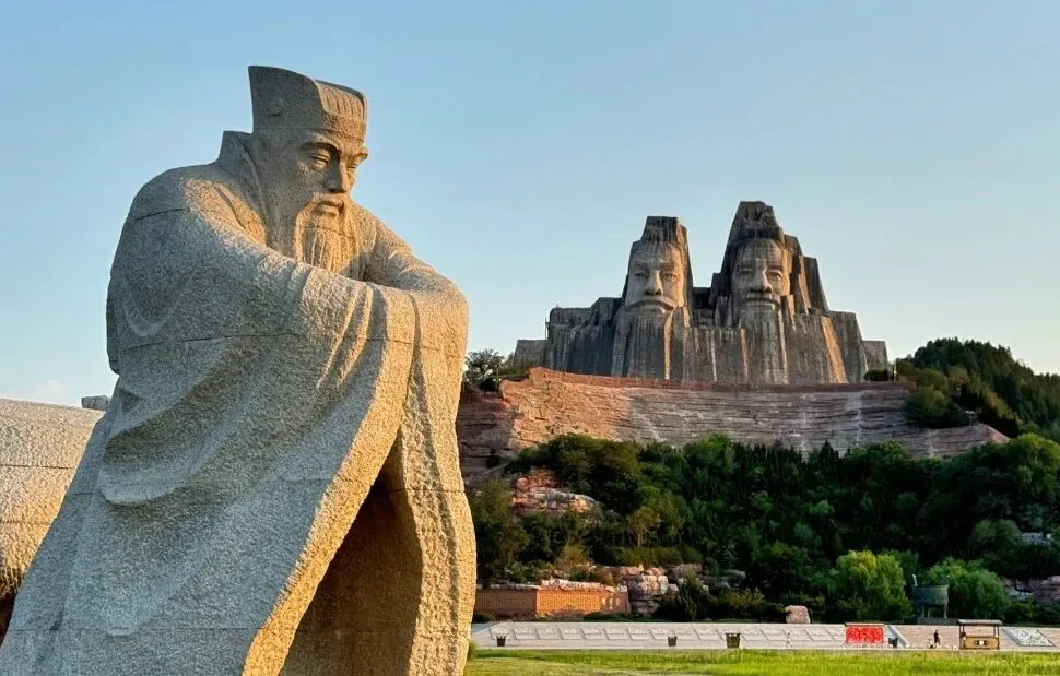 Yellow River Scenic Area,Zhengzhou,Henan,Chinaexplorertour Yellow River Scenic Area,Zhengzhou,Henan,ChinaexplorertourA large seated stone statue in a park with a multi-face rock sculpture carved into a hill in the background, surrounded by trees and landscaping.