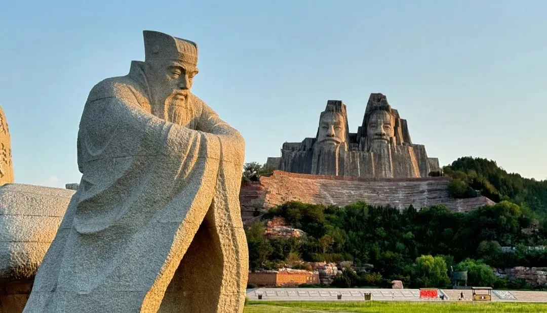 Yellow River Scenic Area,Zhengzhou,Henan,Chinaexplorertour Yellow River Scenic Area,Zhengzhou,Henan,ChinaexplorertourA large seated stone statue in a park with a multi-face rock sculpture carved into a hill in the background, surrounded by trees and landscaping.