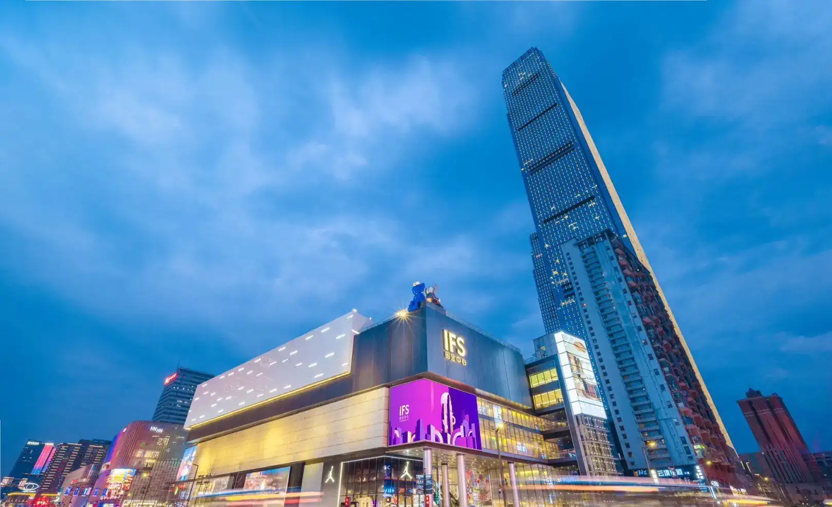IFS International Finance Center Building,Changsha,Hunan,Chinaexplorertour Evening cityscape of a modern mall with bright signage and a tall glass skyscraper against a blue dusk sky.