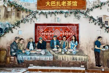 Grand Bazaar,Urumqi,Xinjiang,Chinaexplorertour Mural of a traditional tea-house scene: people dining at two long tables, with a red rug backdrop, greenery, and a sign reading 'Grand Bazaar old Tea House' (Chinese text above).