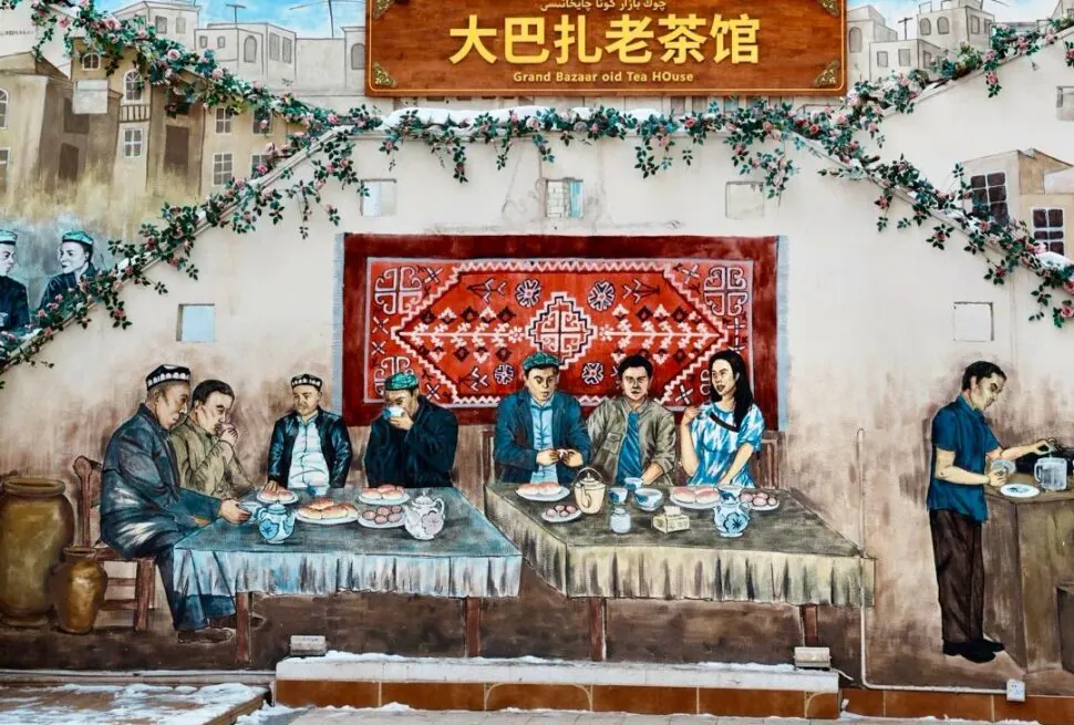Grand Bazaar,Urumqi,Xinjiang,Chinaexplorertour Mural of a traditional tea-house scene: people dining at two long tables, with a red rug backdrop, greenery, and a sign reading 'Grand Bazaar old Tea House' (Chinese text above).