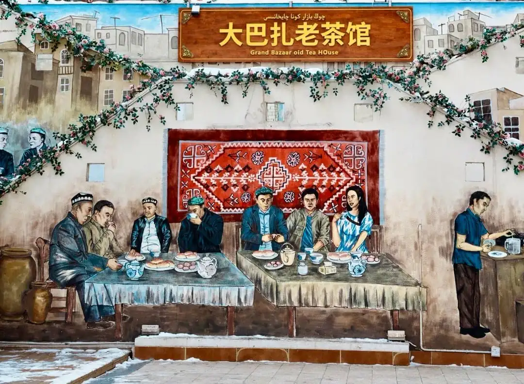 Grand Bazaar,Urumqi,Xinjiang,Chinaexplorertour Mural of a traditional tea-house scene: people dining at two long tables, with a red rug backdrop, greenery, and a sign reading 'Grand Bazaar old Tea House' (Chinese text above).