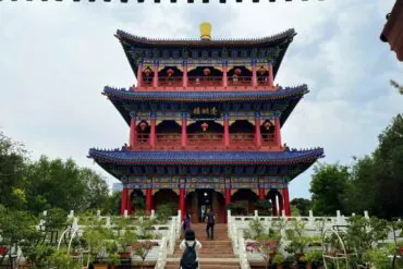 Red Hill Park,Urumqi,Xinjiang,Chinaexplorertour Three-story Chinese temple pavilion with blue roofs, red columns, and red lanterns in a garden front view.