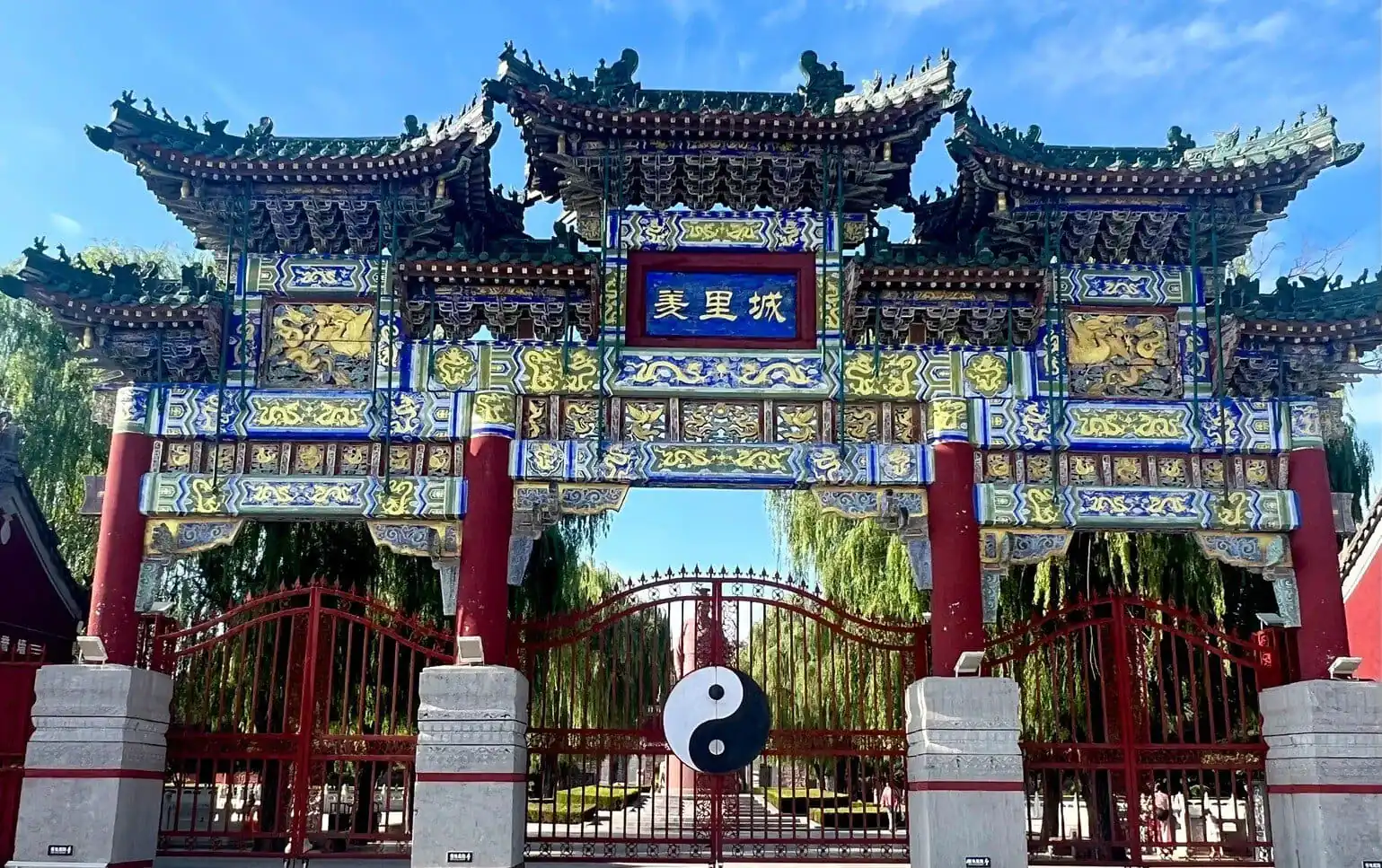 Youli Castle & Scenic Area,Anyang,Henan,Chinaexplorertour Ornate Chinese archway with blue plaque reading '美里城', intricate gold carvings, red pillars, and a yin-yang symbol hanging in front of a gated courtyard.