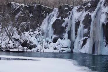 Changbai Waterfall,Baishan,Jilin,Chinatravel Winter scene of a snow-covered cliff with several frozen waterfalls cascading into a blue lake, a bare-branched forest on the left, and a wooden boardwalk along the shore.