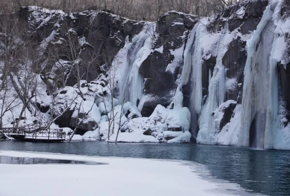 Changbai Waterfall,Baishan,Jilin,Chinatravel Winter scene of a snow-covered cliff with several frozen waterfalls cascading into a blue lake, a bare-branched forest on the left, and a wooden boardwalk along the shore.