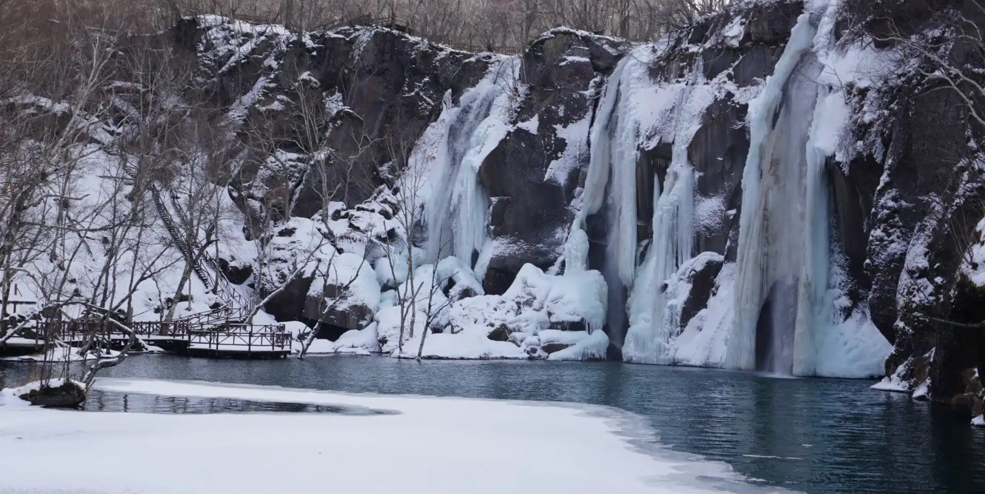 Changbai Waterfall,Baishan,Jilin,Chinatravel Winter scene of a snow-covered cliff with several frozen waterfalls cascading into a blue lake, a bare-branched forest on the left, and a wooden boardwalk along the shore.