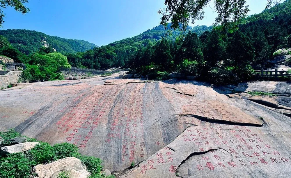 Ancient Chinese rock with red inscriptions surrounded by green trees and mountains.
