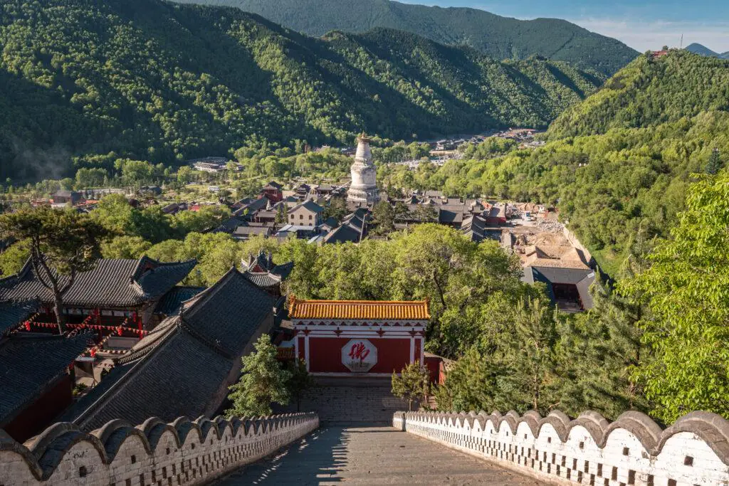 Wide view of a temple complex nestled in green mountains, with a long stone staircase descending toward red gatehouses and traditional roofs.