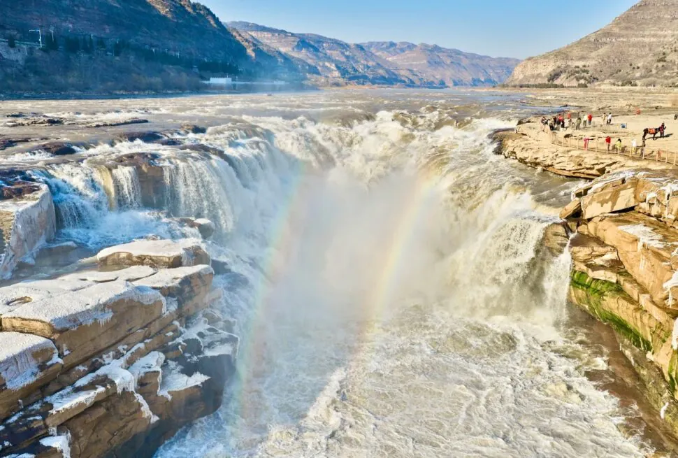 Wide waterfall with mist and a small rainbow, rocky cliffs, and people on a viewing platform to the right,Hukou Waterfall,Linfen,Shanxi