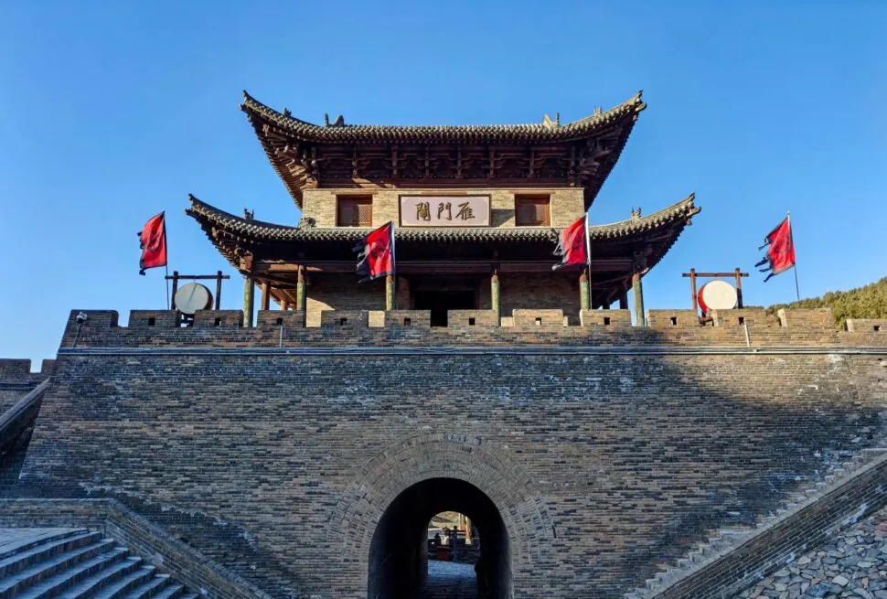 Ancient Chinese city gate with a curved tiled roof and brick walls, red banners on the battlements, and a stone archway beneath a clear blue sky,Yanmen Pass,Xinzhou,Shanxi