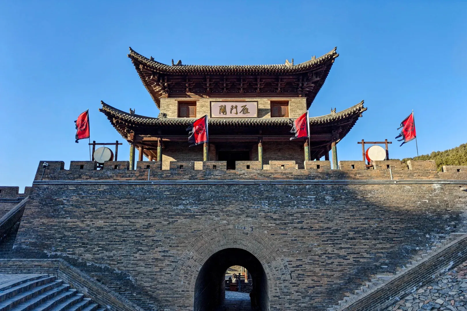 Ancient Chinese city gate with a curved tiled roof and brick walls, red banners on the battlements, and a stone archway beneath a clear blue sky,Yanmen Pass,Xinzhou,Shanxi