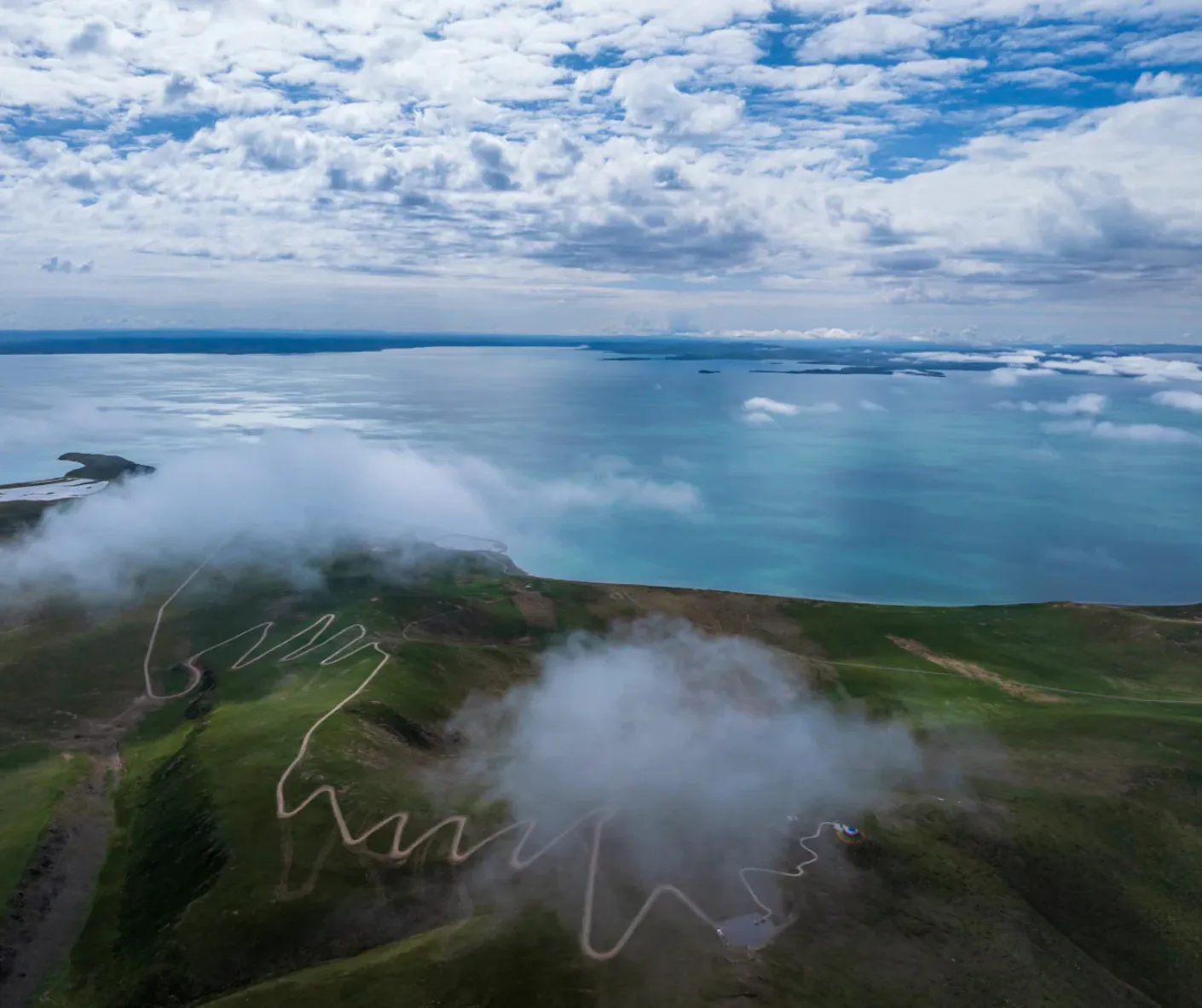 Aerial view of turquoise sea along green hills, with a winding road and cloudy sky above the coastline. Better alt text would be concise and informative for screen readers.Sanjiangyuan National Park,Yushu, Qinghai