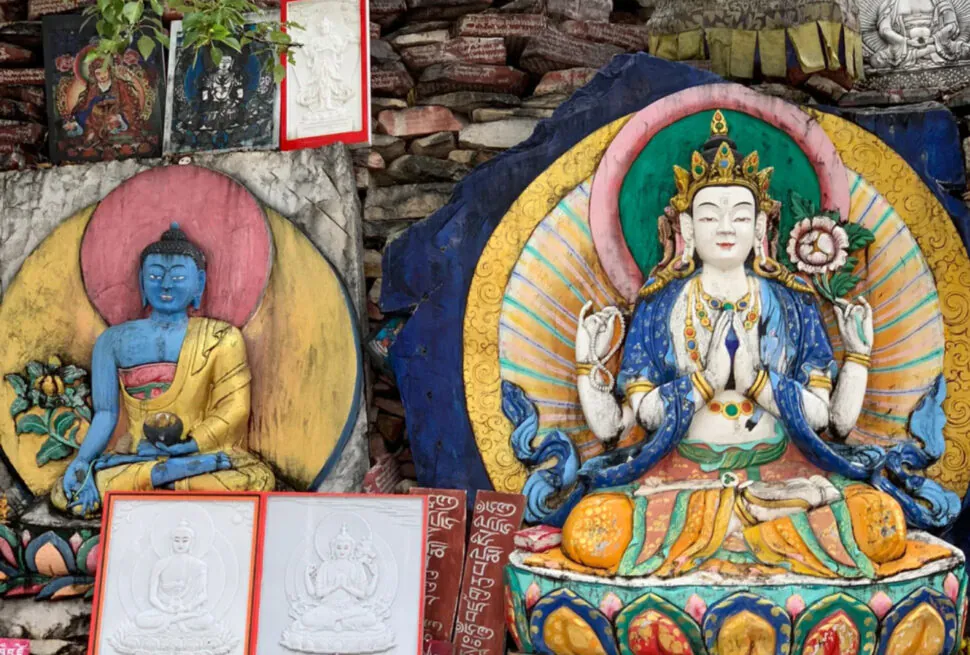 Colorful Buddhist statues and drawings arranged against a stone wall: a large seated multi‑armed deity in the center, a blue Buddha figure on the left, and framed sketches resting on the ground with prayer text visible beneath.Gyanak Mani Stone Mound,Yushu, Qinghai