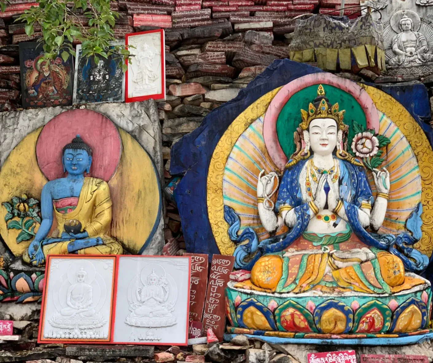 Colorful Buddhist statues and drawings arranged against a stone wall: a large seated multi‑armed deity in the center, a blue Buddha figure on the left, and framed sketches resting on the ground with prayer text visible beneath.Gyanak Mani Stone Mound,Yushu, Qinghai