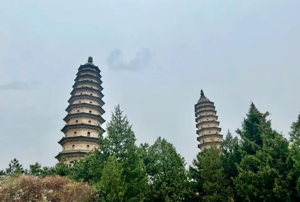 Two tiered stone pagodas rise among green trees against a pale blue sky, creating a tranquil scene.”Shuangta Temple,Taiyuan, Shanxi