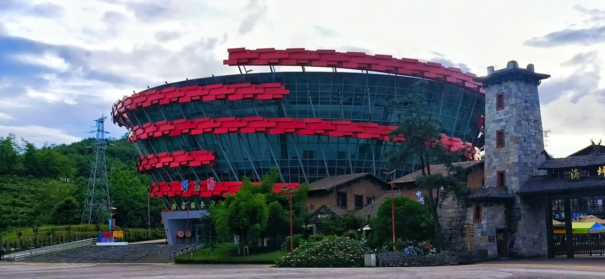 Futuristic circular glass building wrapped in red horizontal panels beside a stone tower and greenery.Yeyuhai Tourist Resort,Liupanshui,Guizhou