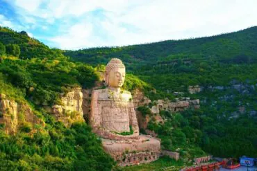 Massive stone Buddha statue carved into a rocky hillside surrounded by dense green foliage and cliffs under a blue sky, overlooking a valley.Mengshan Giant Buddha,Taiyuan, Shanxi