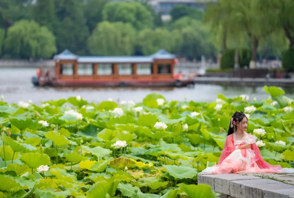 A woman in traditional Chinese dress sitting by a pond with lotus flowers and a boat in the distance.