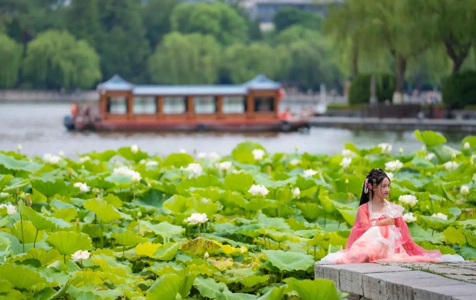 A woman in traditional Chinese dress sitting by a pond with lotus flowers and a boat in the distance.